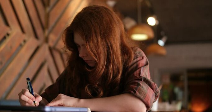 teenage girl with natural red hair and Scottish appearance sits at a cafe table, writing in a notebook. A closed laptop rests on the table. She writes with a fountain pen. Hobby, education