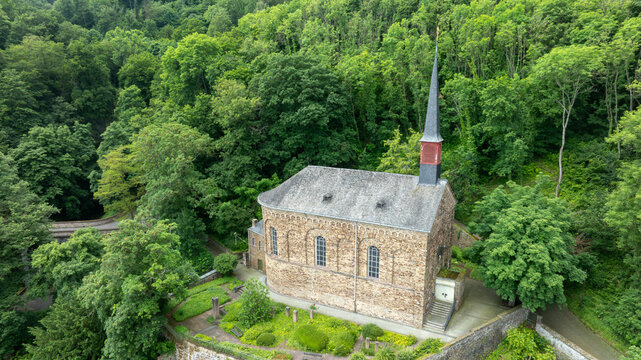 Aerial view of St. Menas church with its tall spire and stone walls nestled within a lush green forest on a hillside in Koblenz, Rheinland-Pfalz, Germany.