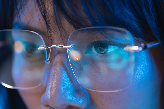 Extreme macro close up of a young Asian woman eye behind a modern rimless eyeglass lens with cool blue light highlighting the iris and her smooth skin texture with a soft cool blue