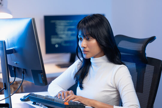 Young Asian woman software developer in elegant white turtleneck typing on her mechanical keyboard at a modern workstation with dual monitors displaying code at night