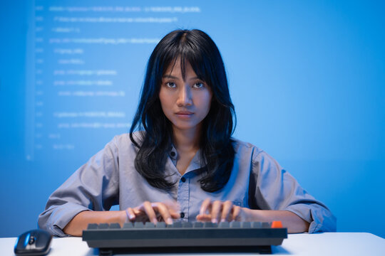 Confident young Asian woman software developer in a blue shirt focused on typing program code on her mechanical keyboard with bright lines of code projected on the wall.