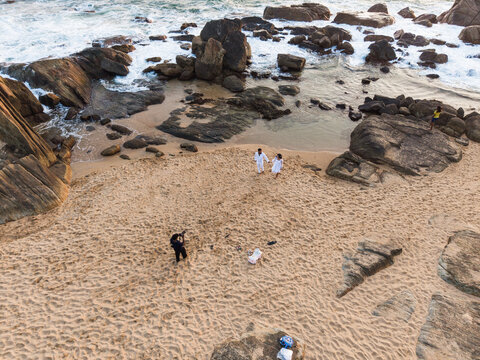 Aerial view of a couple walking on a sandy beach surrounded by large rocks and crashing waves during golden hour in Galle, Southern Province, Sri Lanka.