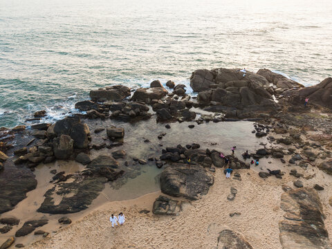 Aerial view of people walking on a sandy beach and rocky shoreline with tide pools and ocean waves in Galle, Southern Province, Sri Lanka.
