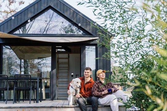 Happy travelers enjoying coffee break outside Scandinavian tiny house with dog