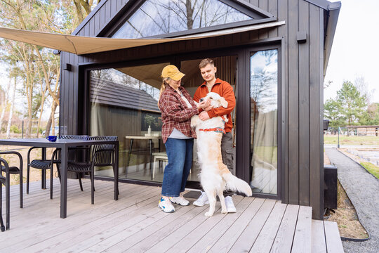Happy couple playing with golden retriever outside modern Scandinavian forest cabin