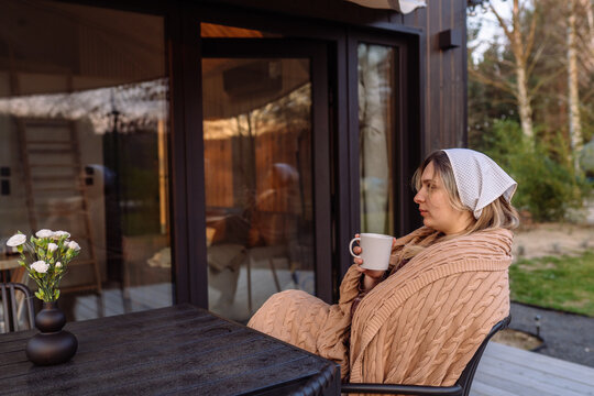 Thoughtful young woman relaxing with warm drink outside modern Scandinavian tiny house