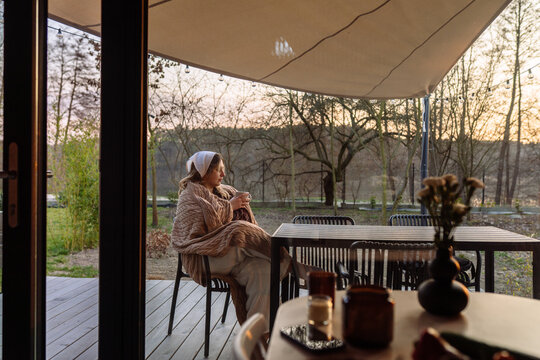 Woman wrapped in knitted blanket drinking tea during quiet sunset cabin retreat