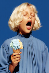 Naklejka premium Blonde child with eyes closed and wide open mouth holding a double scoop blue and vanilla ice cream waffle cone wearing a blue dress against a vivid cobalt background