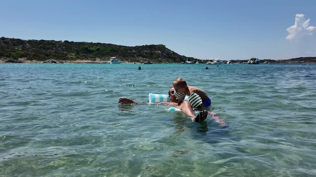 Wide shot of a little boy in armbands jumping into the clear sea water from an inflatable mattress where his father is resting. Beautiful tropical bay, active summer vacation concept.