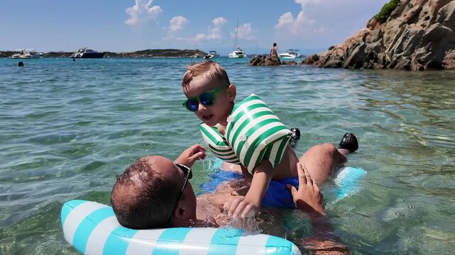 Father lying on a striped inflatable water mattress and holding his little son in armbands, enjoying gentle sea waves during vacation.