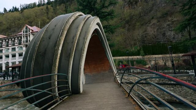 Architecturally striking covered bridge with a curved design spans a river in Borjomi, Georgia, leading towards a quaint building nestled in the mountainside. Path for pedestrians through scenic na...