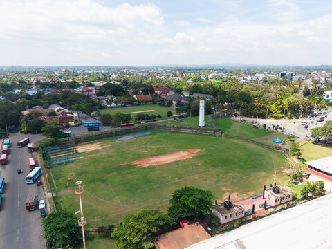 Aerial view of the historic white clock tower and a large green cricket field surrounded by urban buildings and lush trees in Matara, Southern Province, Sri Lanka.
