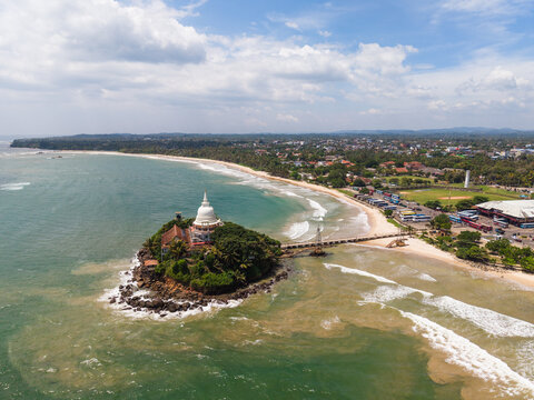 Aerial view of the Parevi Duwa Temple on a small island connected by a bridge to the sandy beach and coastal town in Matara, Southern Province, Sri Lanka.