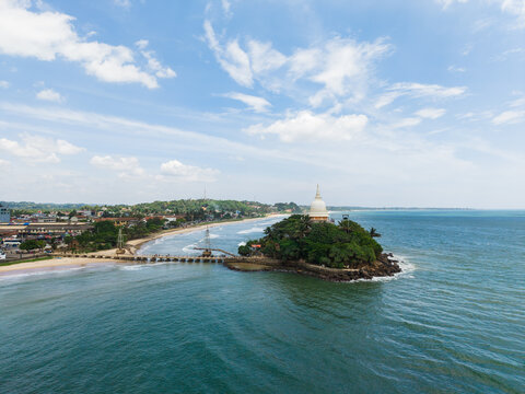 Aerial view of Parevi Duwa Temple on a small island connected by a bridge to the sandy beach and coastal town under a blue sky Matara, Southern Province, Sri Lanka.