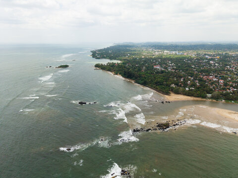 Aerial view of the coastline with waves crashing on rocks and lush green vegetation along the sandy beach in Matara, Southern Province, Sri Lanka.