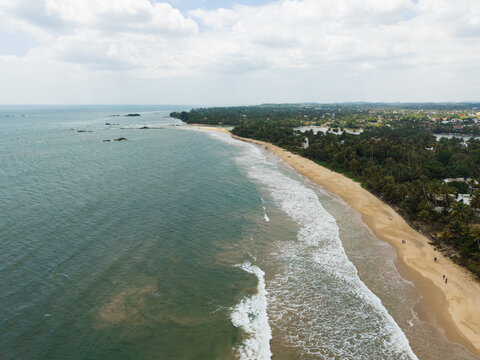 Aerial view of a long sandy coastline with turquoise waves crashing on the shore and lush tropical palm trees in Matara, Southern Province, Sri Lanka.