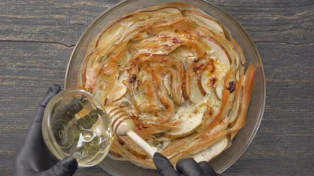 Top view of honey pouring onto a baked phyllo dough pie with pears and cheese. Sweet syrup drizzling over crispy pastry on a table