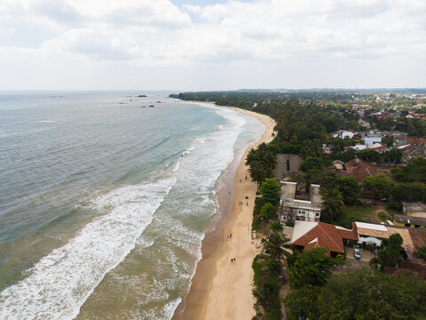 Aerial view of the sandy coastline with turquoise waves crashing on the shore, palm trees, and coastal buildings Matara, Southern Province, Sri Lanka.