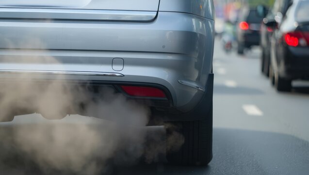 Extreme close-up of a silver car's rear bumper with thick exhaust fumes billowing from the tailpipe while idling in an urban traffic queue, highlighting vehicle emissions and pollution.