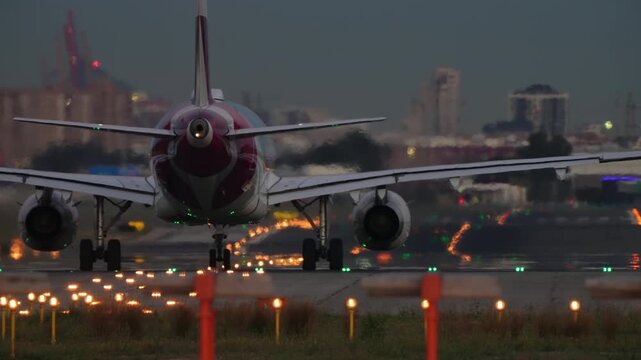Commercial aircraft with red livery rolls on runway at dusk, glowing runway lights creating colorful orange, green and yellow bokeh patterns across the wet reflective tarmac.
