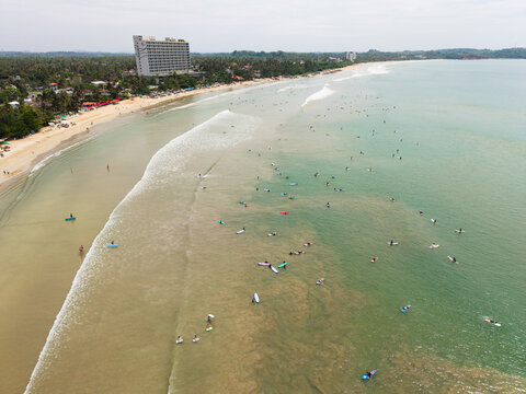 Aerial view of surfers catching waves along the sandy shoreline and tropical palm trees Weligama, Southern Province, Sri Lanka.