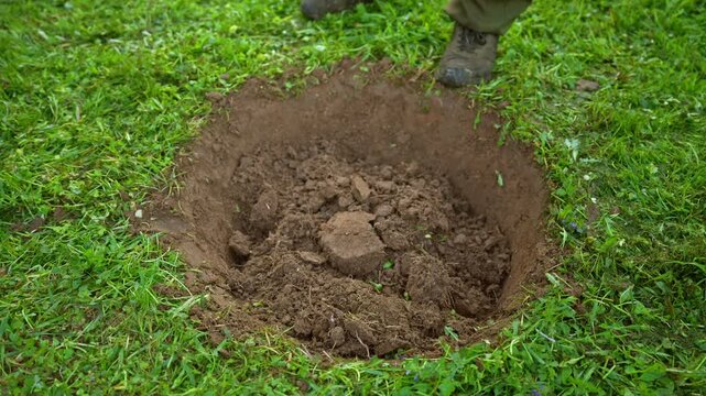 Close up view of a person wearing boots using a shovel to fill a freshly dug pit with earth on a green lawn, representing manual labor, burial, or gardening preparation for planting