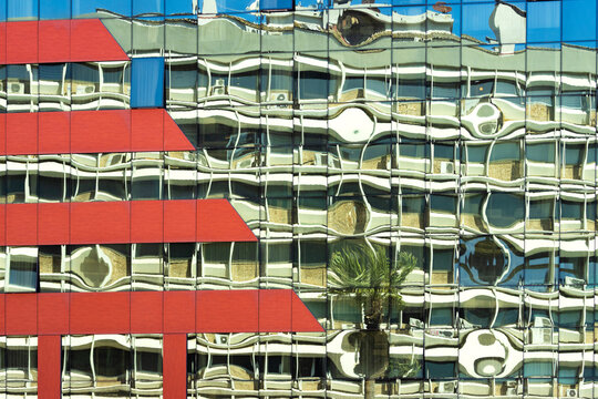 Architectural reflection concept: modern blue glass skyscraper facade with red panels reflecting an old building structure and a palm tree.