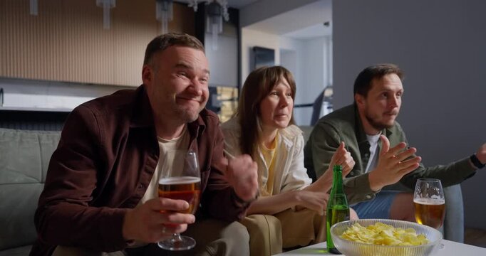Three friends gathered on a comfortable couch in a modern living room enjoying drinks together, watching something intently and celebrating joyfully, surrounded by snacks and a relaxed atmosphere