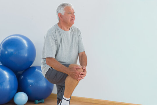Senior man in gray t-shirt performing knee hug in therapy room with blue exercise balls, dumbbell