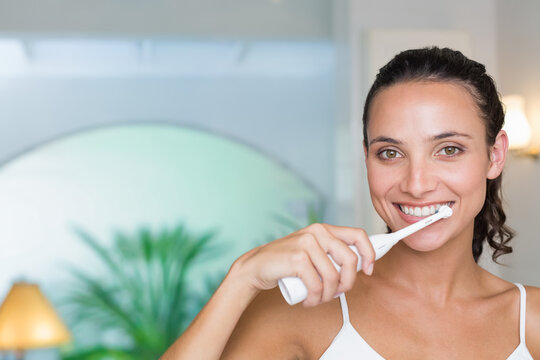20s-woman standing at bathroom vanity brushing teeth with electric toothbrush in tank, copy space