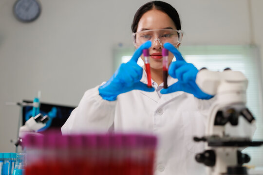 Female scientist wearing safety glasses and blue gloves, holding and examining two blood sample tubes, performing research and analysis in a professional laboratory setting