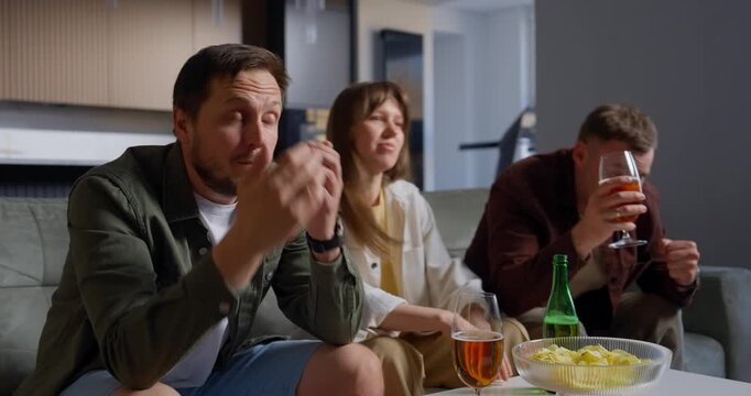 Three friends sitting on a couch in a living room, watching something intensely with varying emotions, from focused and tense to smiling, with snacks and drinks on the table in front of them