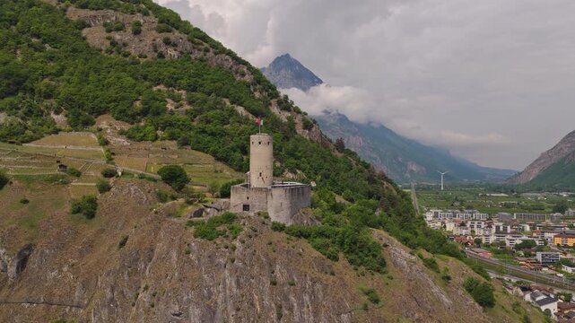 Cinematic drone footage of Ch&acirc;teau de la B&acirc;tiaz in Martigny, Switzerland, featuring the historic medieval tower, surrounding mountains and panoramic views over the alpine town on a sunny bluebird day.