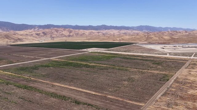 Arid landscape of Central California showing the contrast between dry, fallow fields and a lush, irrigated orchard, with distant mountains and a clear blue sky creating a striking backdrop