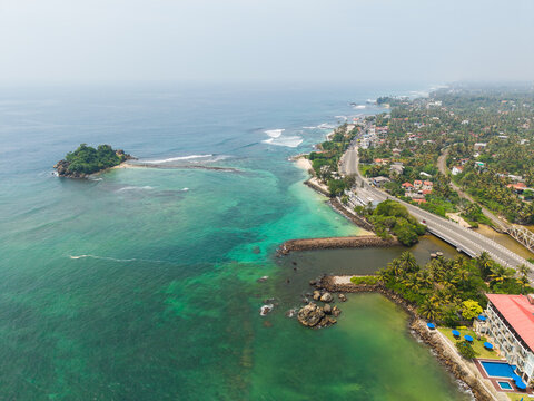 Aerial view of Devil's Rock island surrounded by turquoise ocean waters, a coastal road, and lush tropical greenery at Ahangama, Southern Province, Sri Lanka.