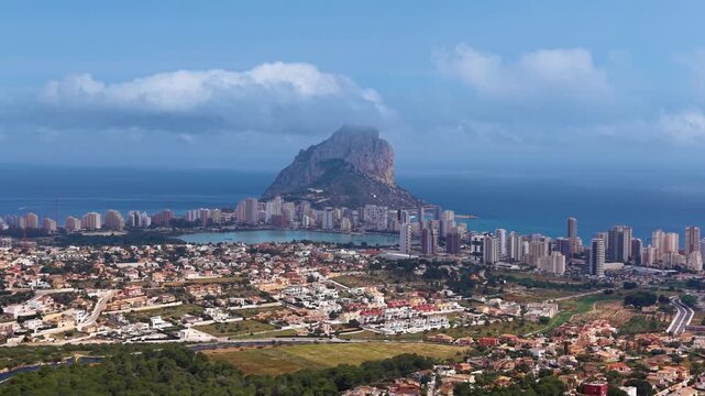 Panoramic view of Calpe, Spain, featuring the iconic Penon de Ifach rock formation rising above the coastal city, with turquoise Mediterranean sea and clouds overhead