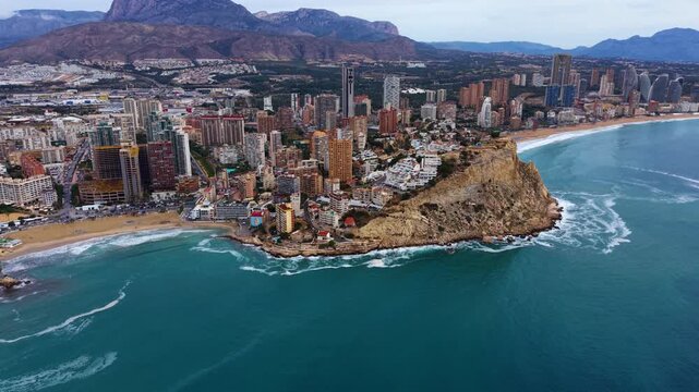 Benidorm Spain coastline with turquoise Mediterranean Sea, rocky headland, sandy beaches, high-rise buildings and mountain backdrop under overcast skies