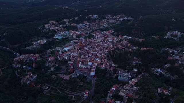 Serra village at dusk in Spain, small hillside town surrounded by dense forest, terracotta rooftops and winding roads visible from above in fading evening light