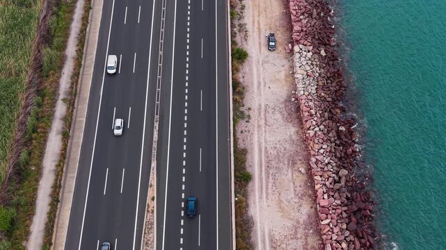 Overhead view of a multi-lane coastal highway with cars driving alongside a rocky shoreline and turquoise sea water