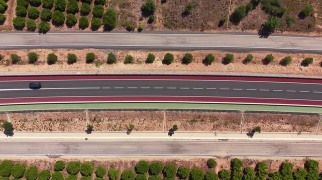 Top-down view of a modern dual carriageway with colorful red and green cycle lanes, a single black car driving, flanked by rows of green trees and dry scrubland