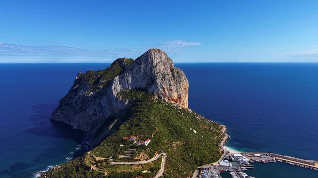 Iconic Penon de Ifach rock formation rises above the Mediterranean Sea in Calpe, Spain, with a marina and lush green vegetation visible at its base