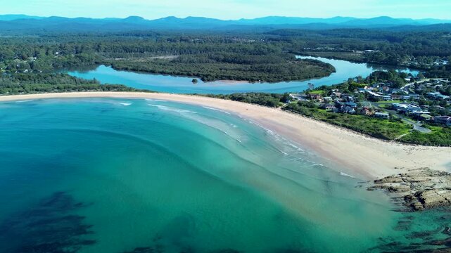 Drone aerial landscape of seaside waves breaking on sandy beach shoreline with Tomaga River inlet surrounded by forest bushland trees in rural town suburbs of Tomakin Eurobodalla NSW Australia tourism