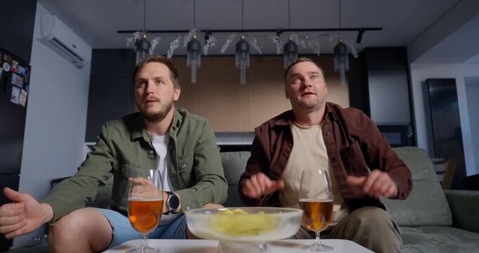 Two young men sitting on a couch in a modern living room, sharing drinks and snacks while watching something engaging on TV, displaying different emotions from focused to happy
