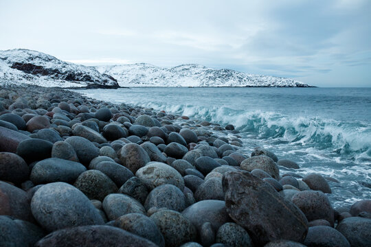 Rocky beach with round stones in winter Teriberka Arctic ocean coast background with snow covered hills and sea waves, Barents sea shore, northern landscape, cold dramatic nature travel concept