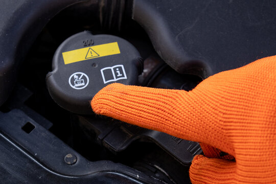 A mechanic uses a gloved hand to point at the antifreeze tank cap on a vehicle while performing maintenance in a garage