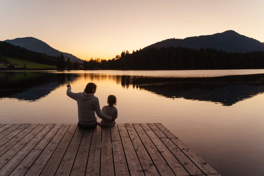 Mother and little daughter sitting together at end of lakeshore jetty at dusk