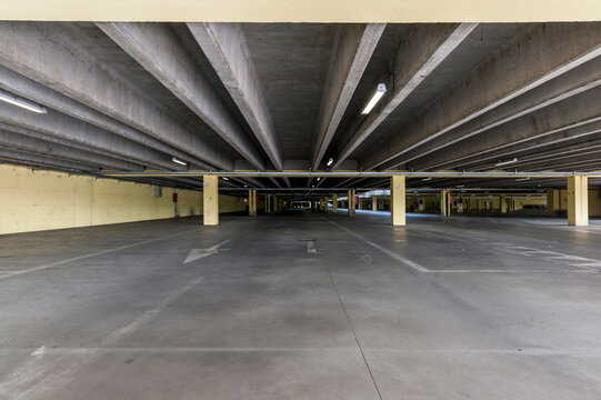 Parcheggio coperto vuoto con pavimento e soffitto in cemento, sostenuto da pilastri beige. L'illuminazione fluorescente illumina l'ampia area. Frecce  bianche e linee di parcheggio sul pavimento.