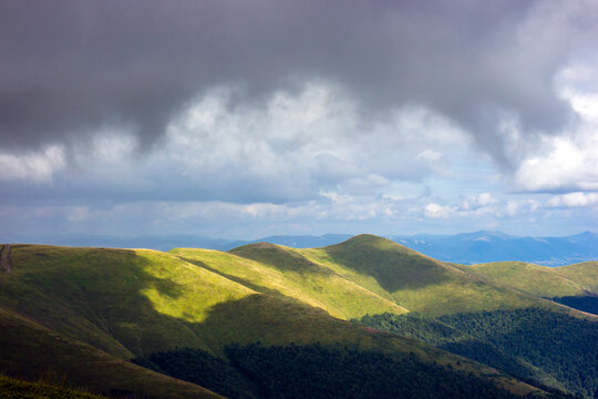 Dramatic storm clouds cast moving shadows over rolling green mountain hills under soft sunlight.