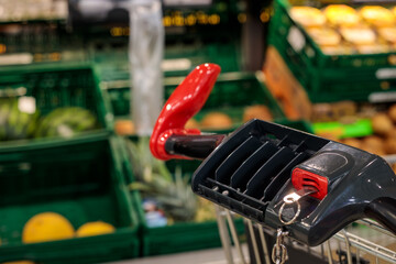 close-up of shopping cart red handle with coin lock and key ring detail, blurred supermarket fruit section with melons and kiwi in background © Татьяна Швец