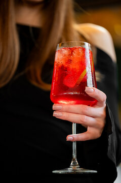 A close-up of a woman holding a large wine glass filled with a refreshing red cocktail, ice cubes, and a slice of citrus against a dark background.